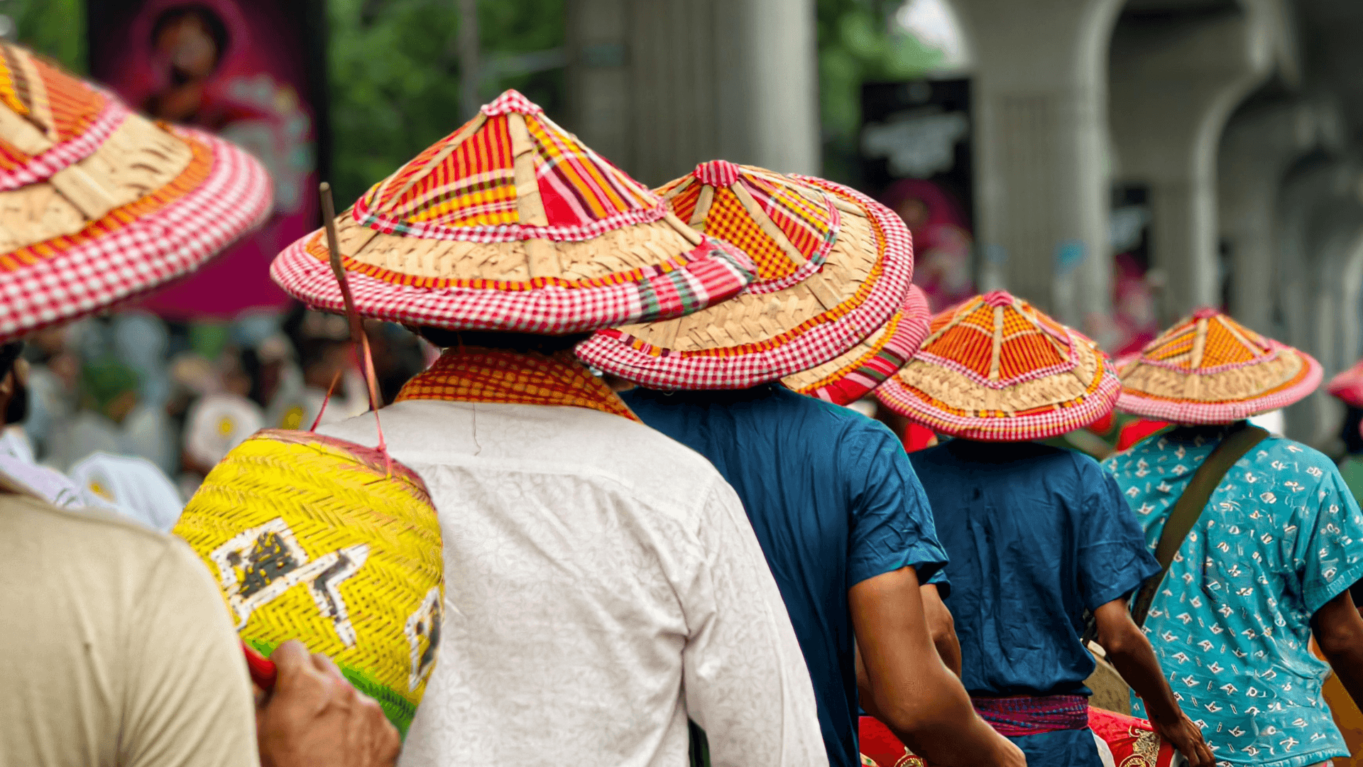 A group of people wearing colorful, traditional woven hats and carrying baskets walk in a line outdoors, with greenery and columns visible in the background.