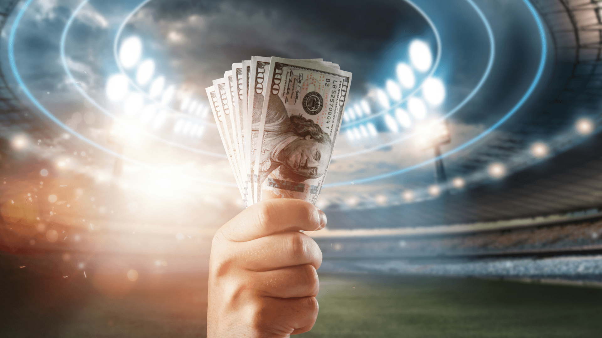A hand holding a fan of U.S. hundred dollar bills in the foreground, with a brightly lit stadium and cheering crowd in the background.