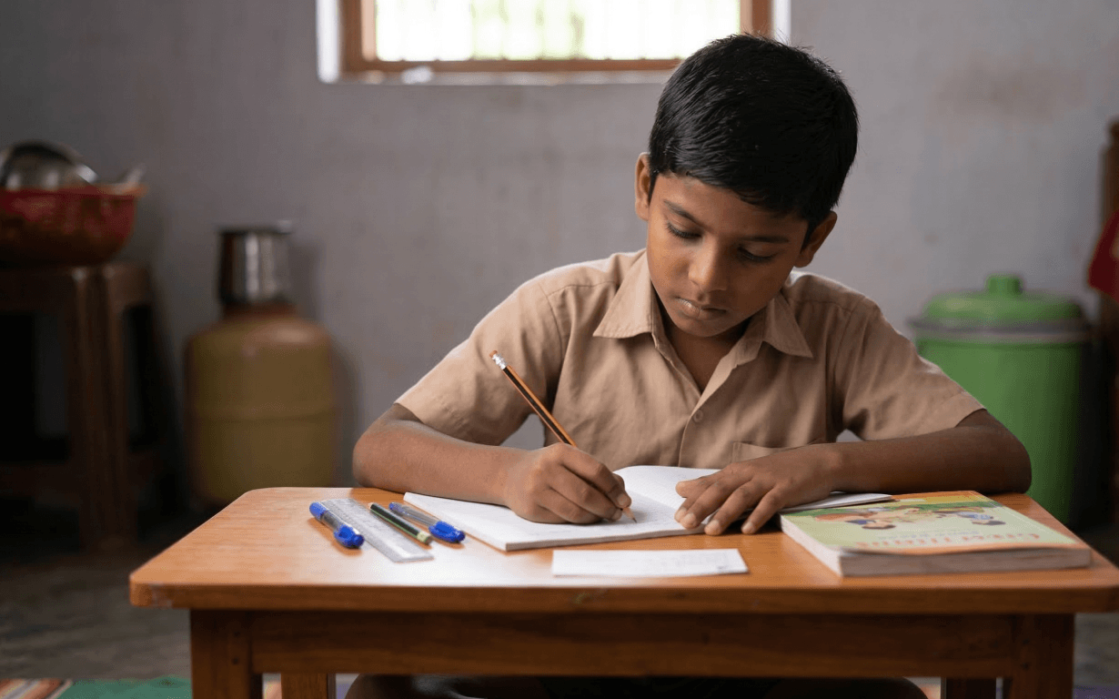 A young boy sits at a wooden desk, focused on writing in a notebook with a pencil. Several pens, a book, and notes about remittances are on the desk. The room has simple furnishings and daylight comes through a window behind him.
