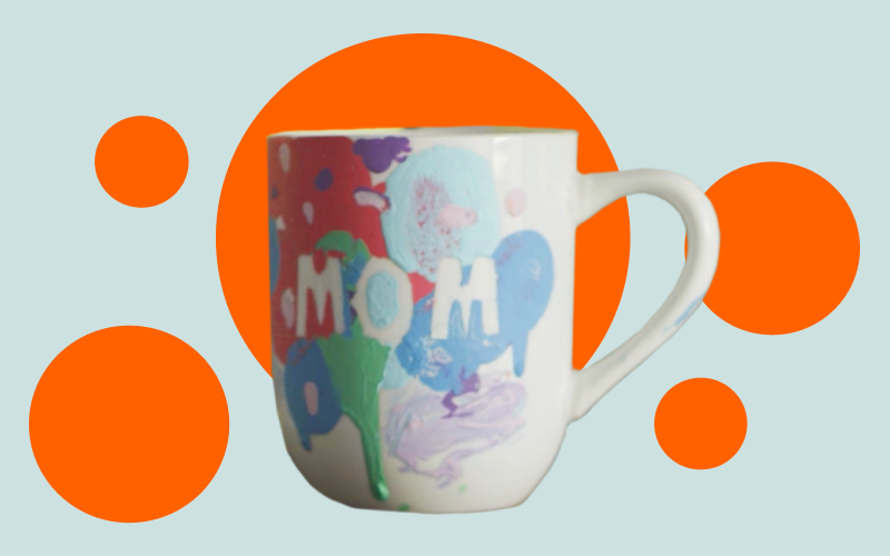 A white ceramic mug decorated with colorful, abstract paint splashes and the word "MOM" in bold white letters. The background features large, bright orange circles on a light blue surface, giving the image a vibrant and playful feel.