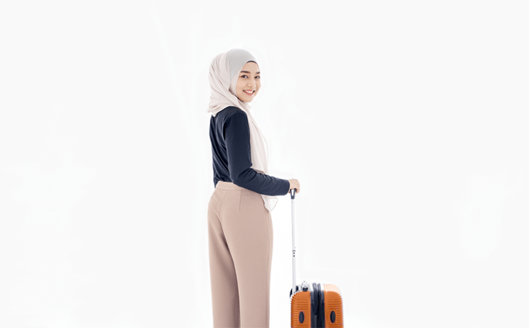 Une personne souriante portant un hijab portant un haut noir à manches longues et un pantalon beige se tient légèrement tournée vers l'arrière, tenant la poignée d'une valise orange sur un fond blanc uni.
