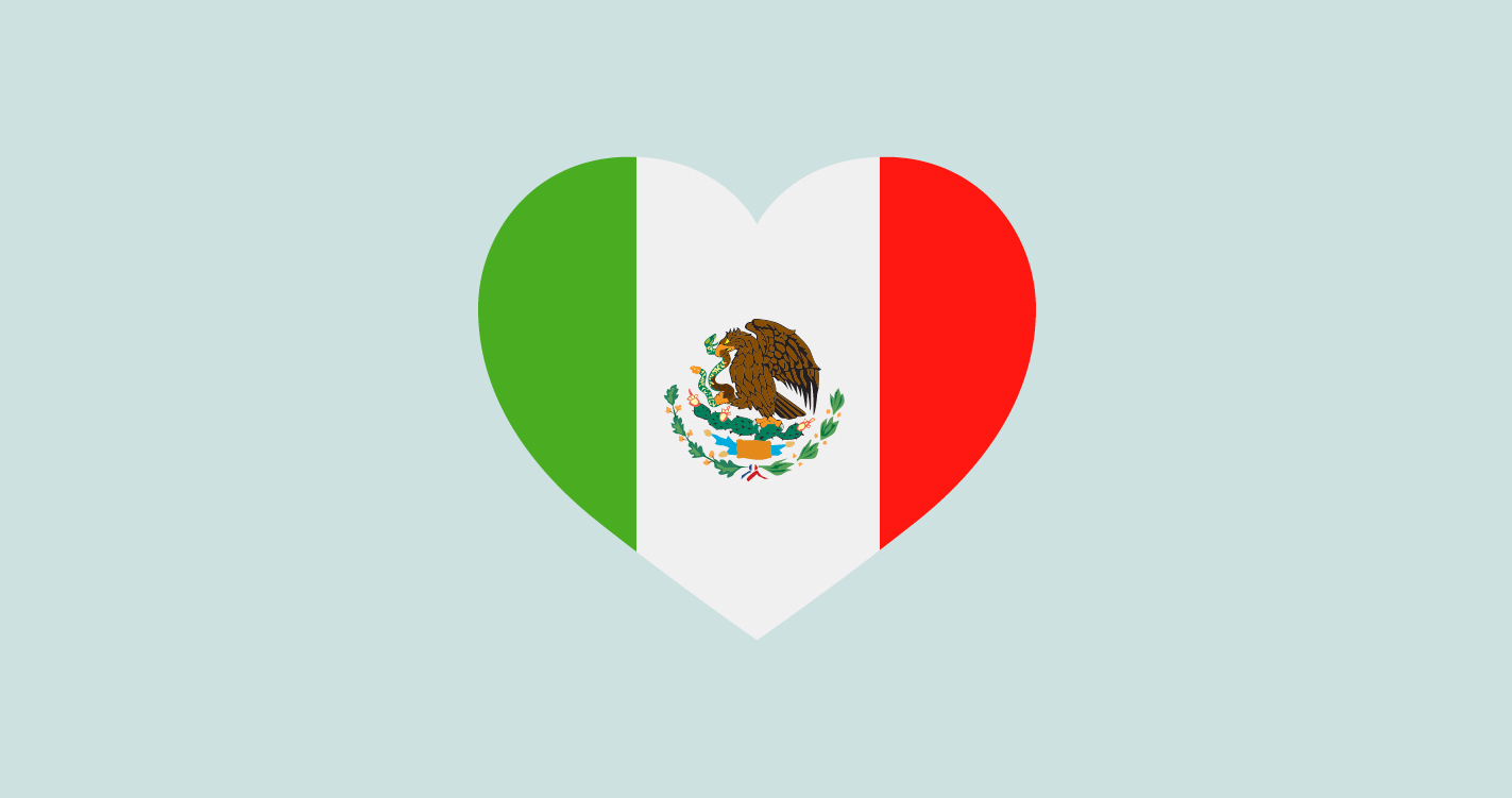 Una forma de corazón llena con la bandera de México sobre un fondo azul claro. El corazón contiene tres franjas verticales de verde, blanco y rojo de izquierda a derecha, con el emblema nacional de México, un águila sosteniendo una serpiente sobre un cactus, centrado en la franja blanca.