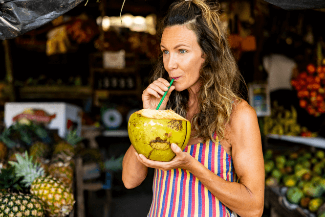 A woman with wavy hair drinks from a fresh coconut using a green straw. She is wearing a striped tank top and standing in a vibrant market stall filled with tropical fruits, including pineapples and mangoes, visible in the background—truly embracing the essence of moving to Costa Rica.