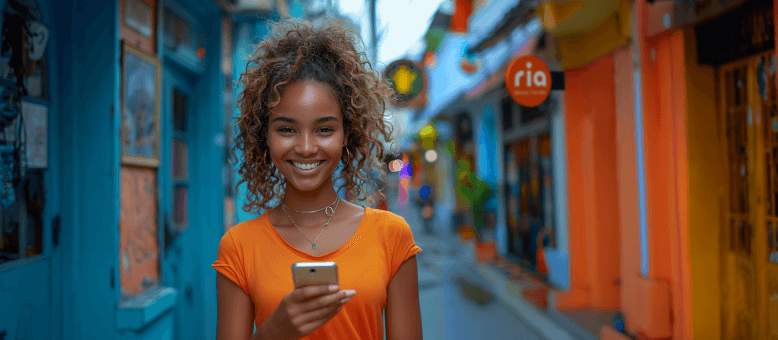 Una mujer sonriente con una camiseta naranja sostiene un teléfono inteligente en una calle vibrante y colorida con varias tiendas y carteles. El fondo está ligeramente borroso y en un cartel se lee "ria". La escena sugiere un ambiente animado y alegre.
