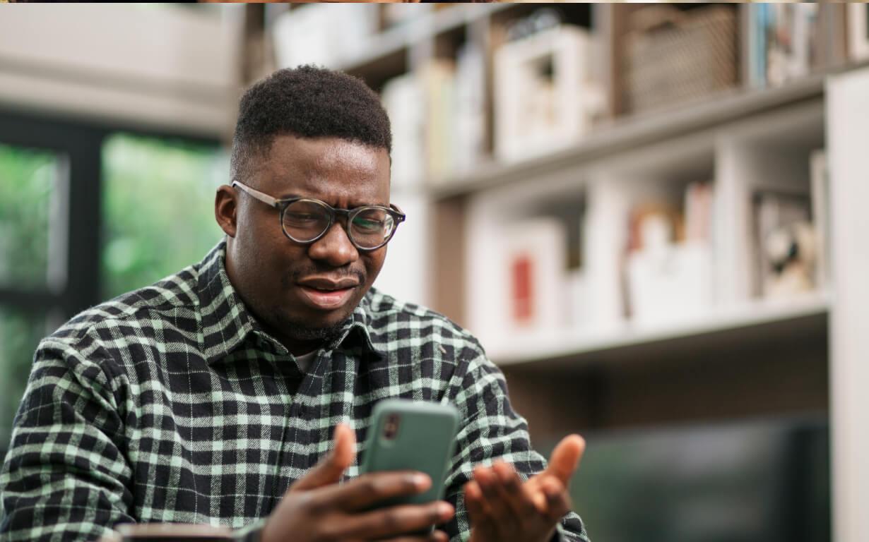 A man wearing glasses and a plaid shirt looks frustrated while holding a smartphone. Indoors, surrounded by shelves and books, he gestures with his other hand, questioning the unpredictable currency exchange rates displayed on the screen.