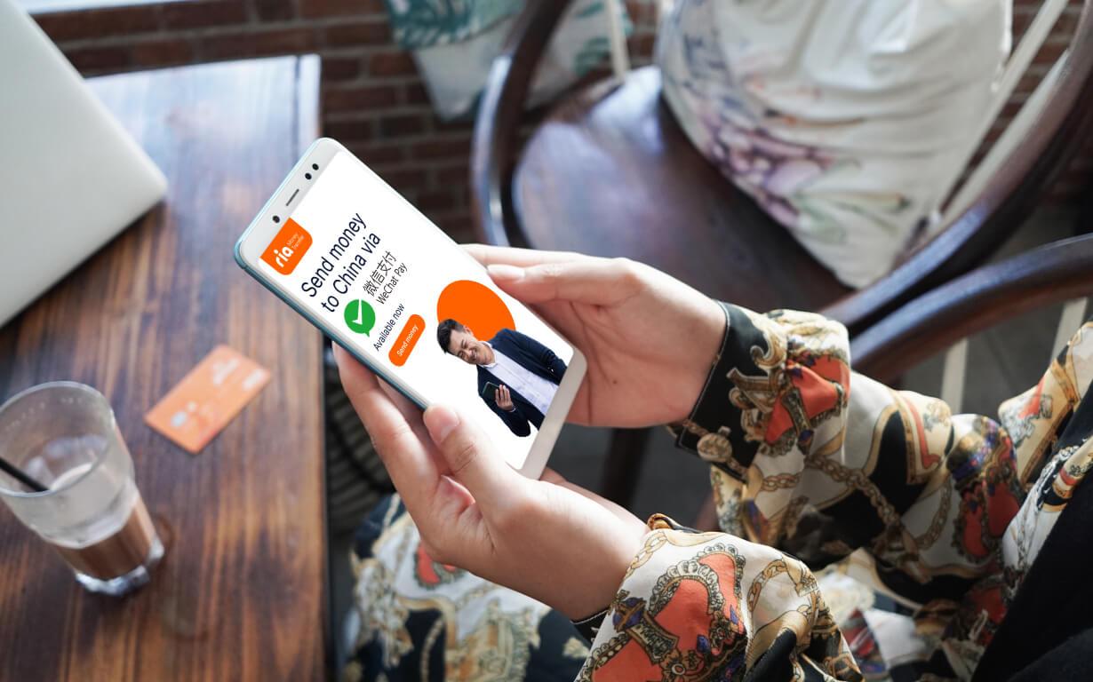 A person is holding a smartphone showing a WeChat money transfer app screen with an image of a businessman, options to "Send now," and a completed transaction message. Nearby, a table holds a drink and card, creating the perfect scene for seamless transactions.