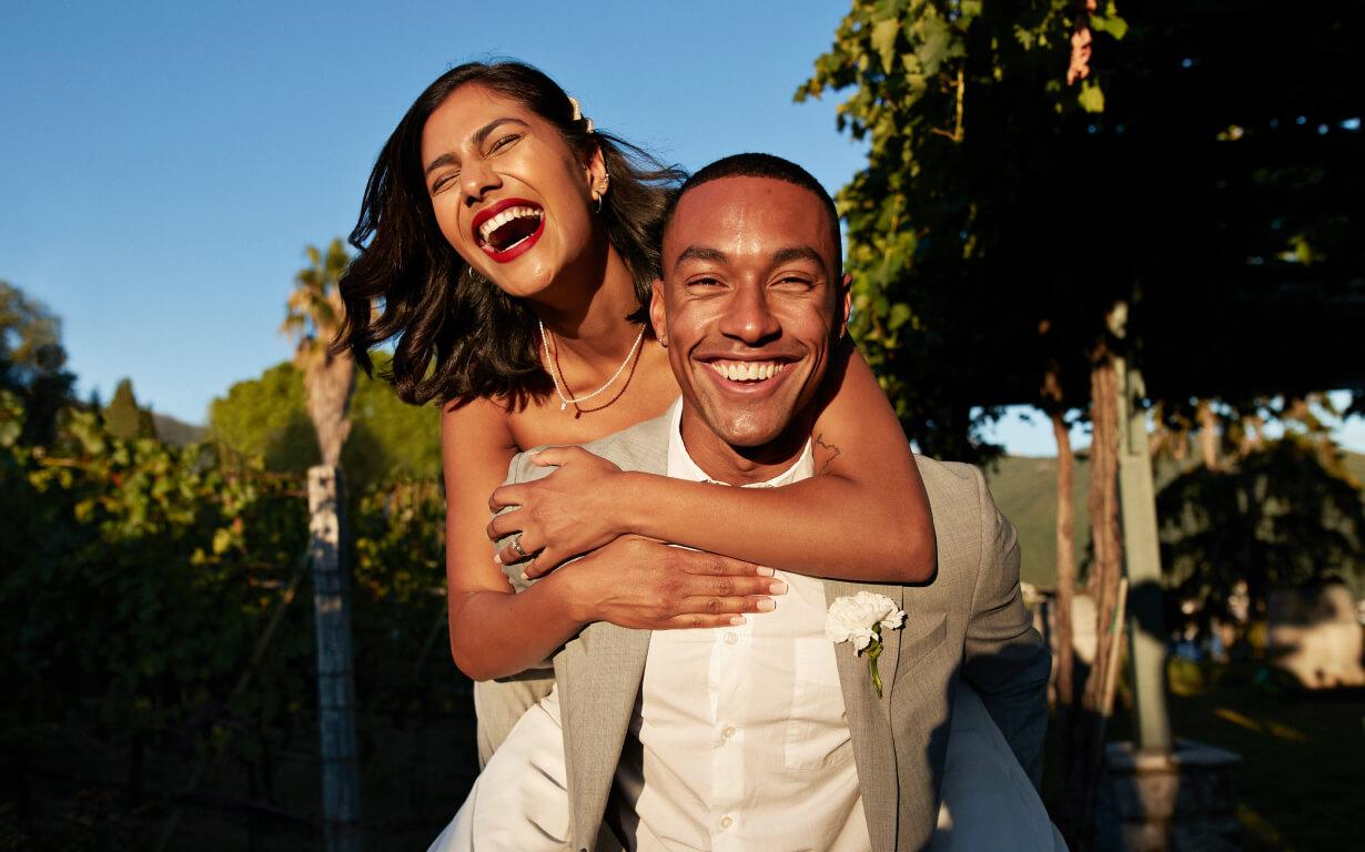 Una pareja alegre en una boda al aire libre. La mujer, con un vestido blanco, se ríe y lleva a caballito al hombre, con un traje gris. Están rodeados de vegetación y luz solar, con árboles y un cielo azul claro de fondo.