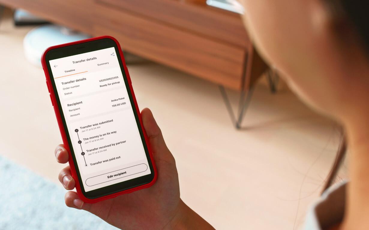A person holds a smartphone displaying bank transfer details, including Track Ria Transfer status updates and recipient information. The softly blurred background draws attention to the phone’s display.
